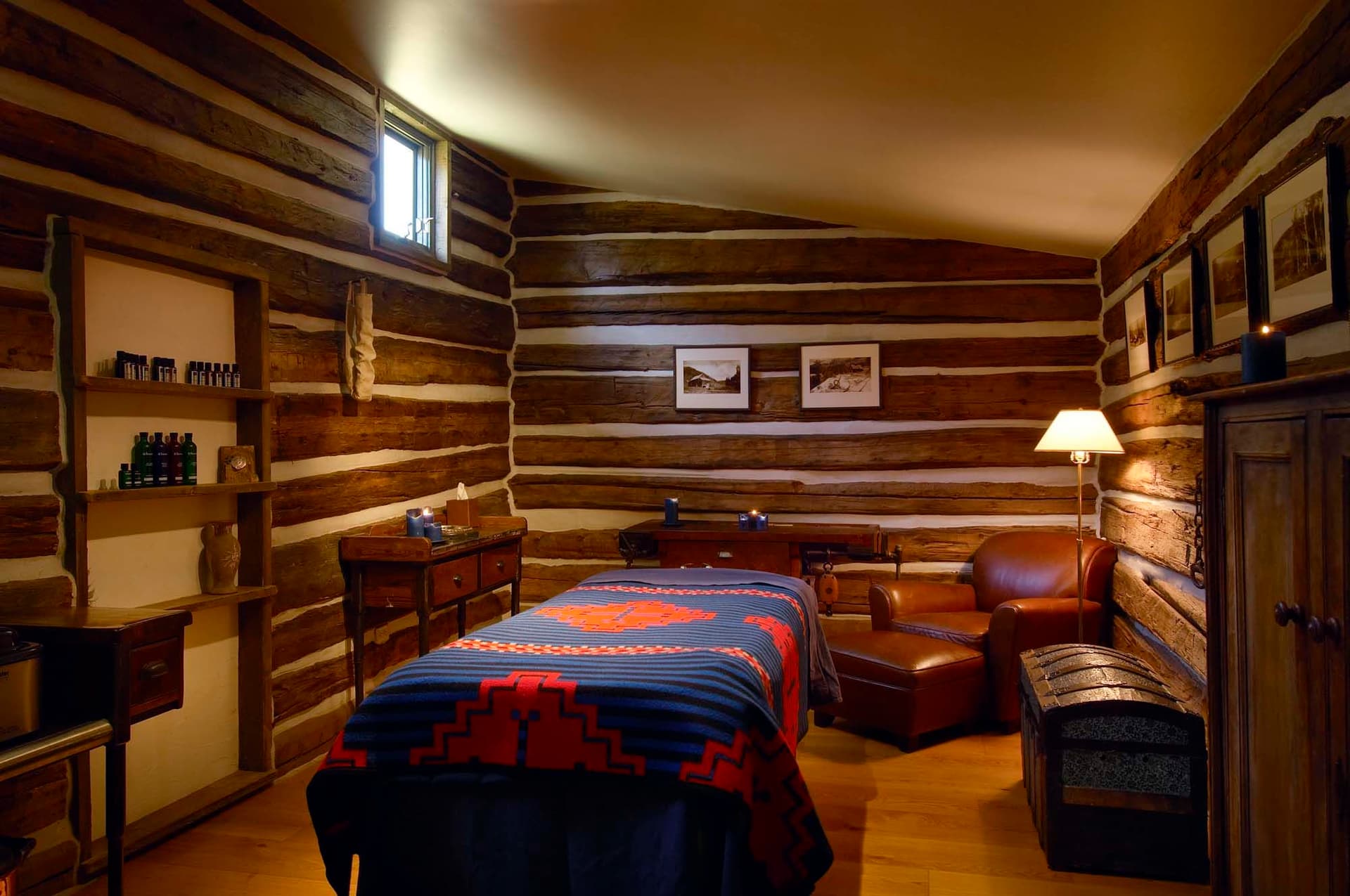 Cozy log cabin room with massage table, warm lighting, wooden walls, framed pictures, and shelves with oils.