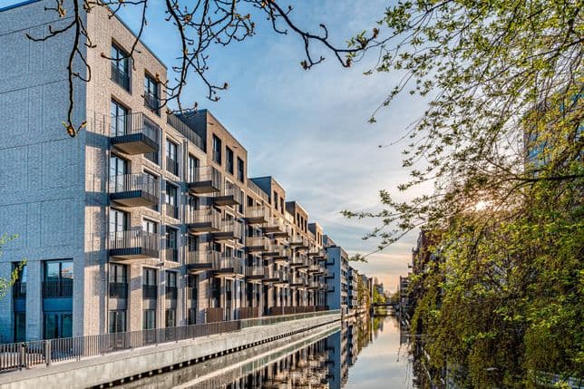 Modern canal-side apartment buildings with balconies, reflecting in the water, surrounded by trees under a clear blue sky at sunset.