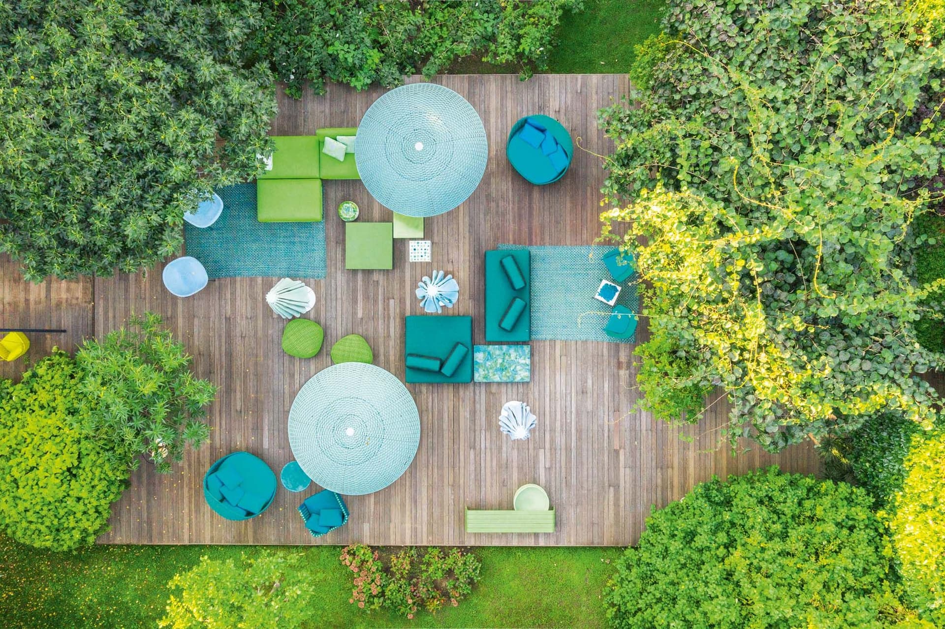 Aerial view of a wooden terrace with green and blue garden furniture and large parasols, surrounded by lush greenery.