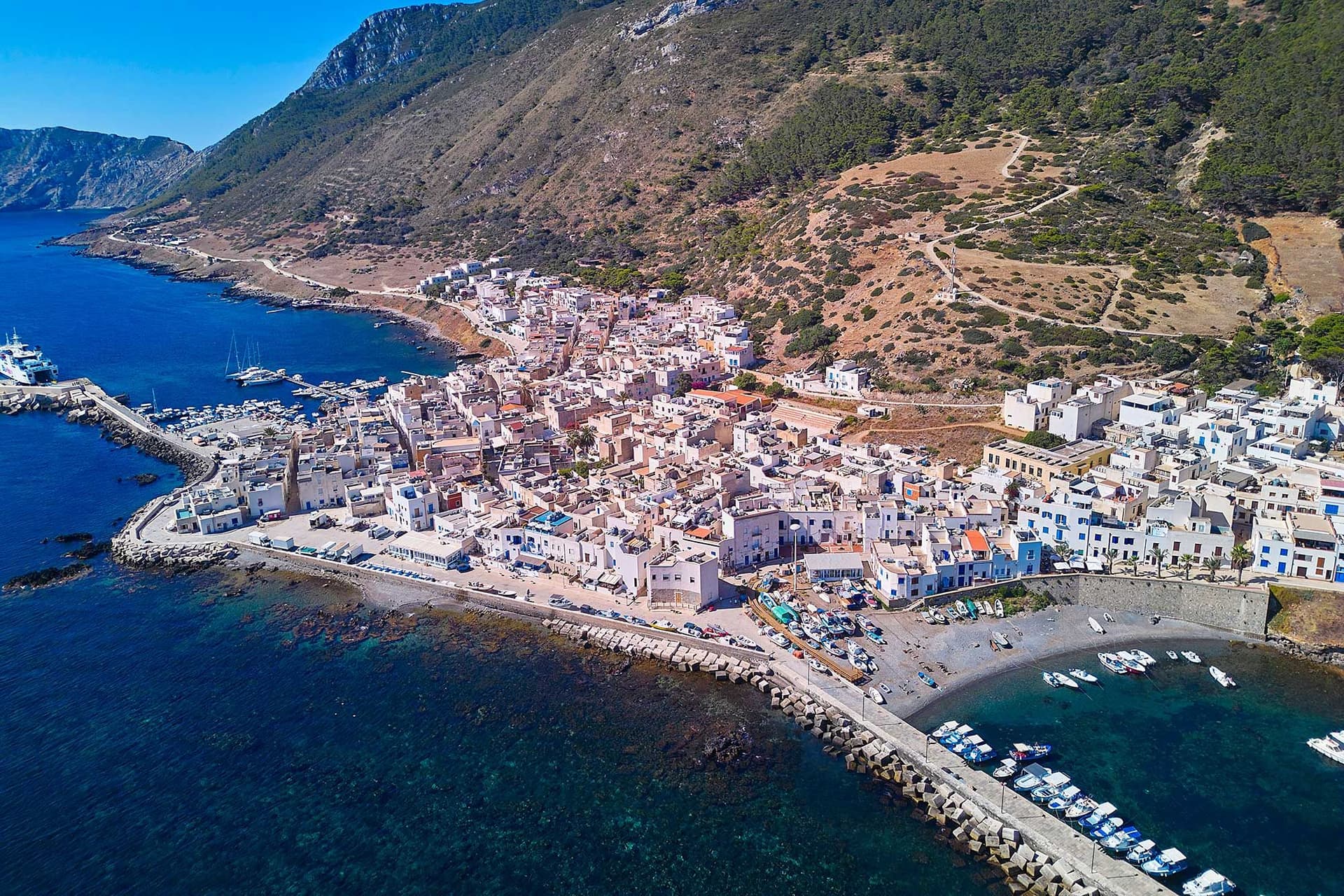 Veduta aerea di un villaggio costiero con edifici bianchi, un porto turistico e le colline circostanti in riva al mare, sotto un cielo azzurro e limpido.