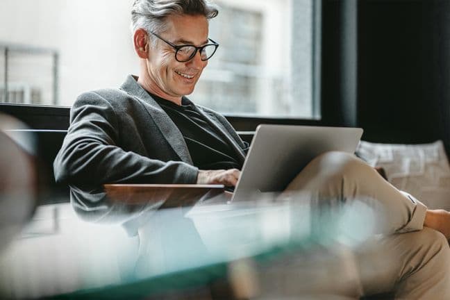 A mature businessman wearing glasses, a grey blazer, and black shirt sits comfortably on a couch, smiling while working on his laptop. The setting appears to be a bright, modern home office space.