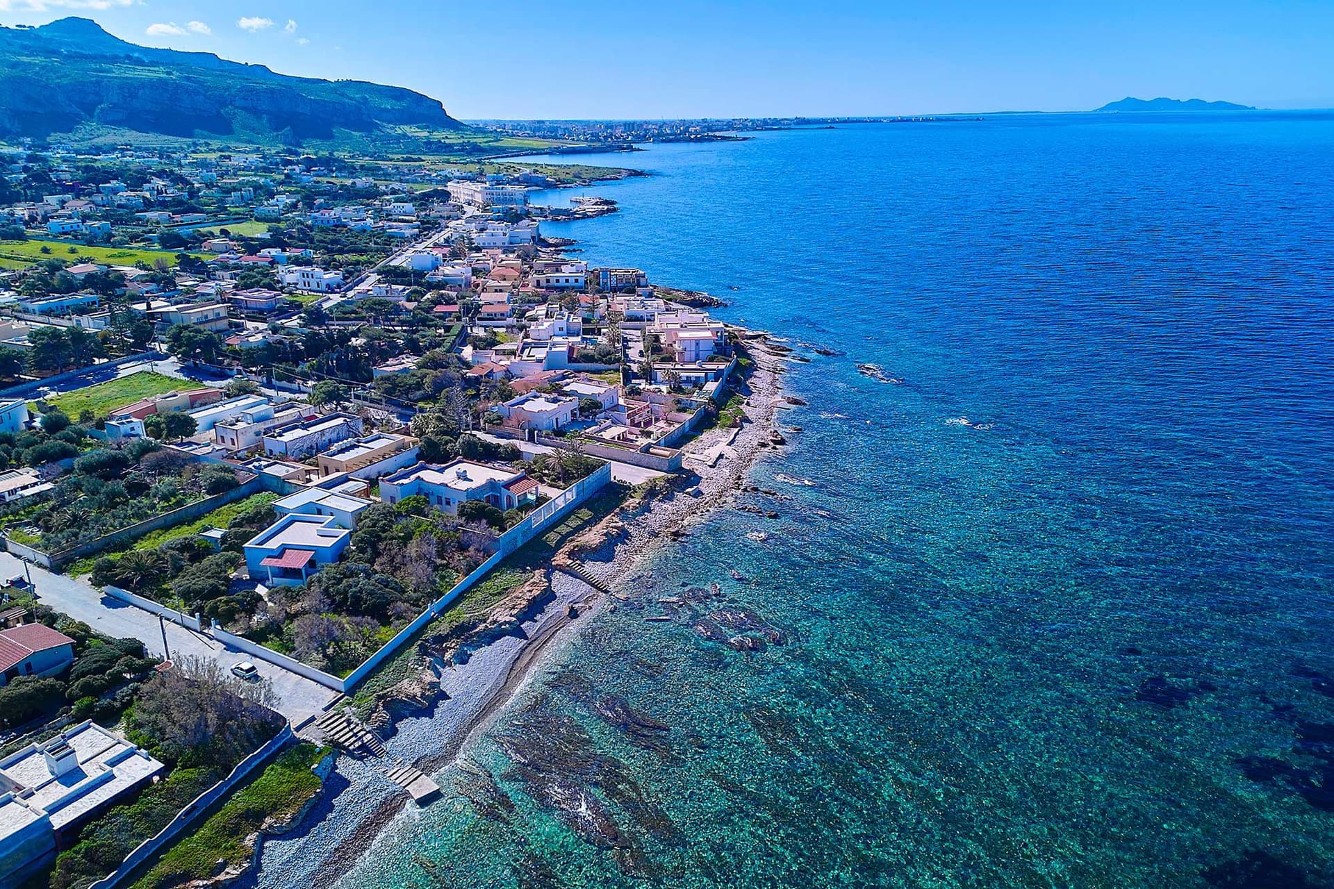 Aerial view of a coastal town with clear blue sea, rocky shoreline, and houses surrounded by greenery under a bright blue sky.