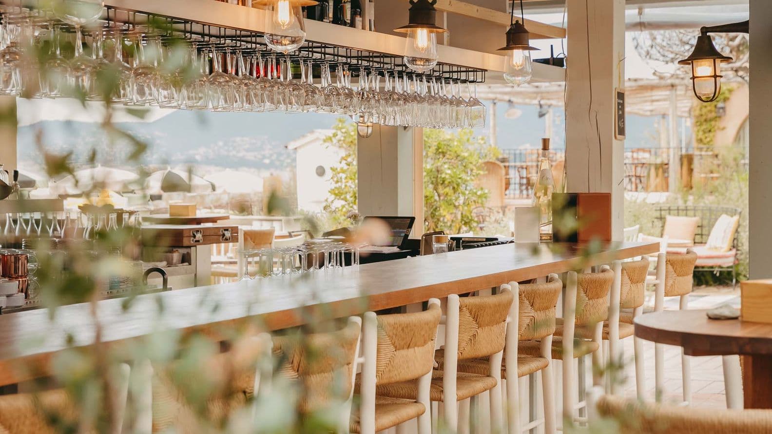 Cozy outdoor bar with wooden stools and hanging glasses, surrounded by greenery, offering a view of a distant landscape.