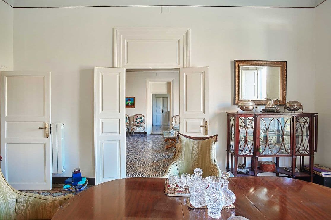 Elegant dining room with a wooden table, crystal decanters, and a glass cabinet. Open doors reveal a patterned floor and additional seating area.