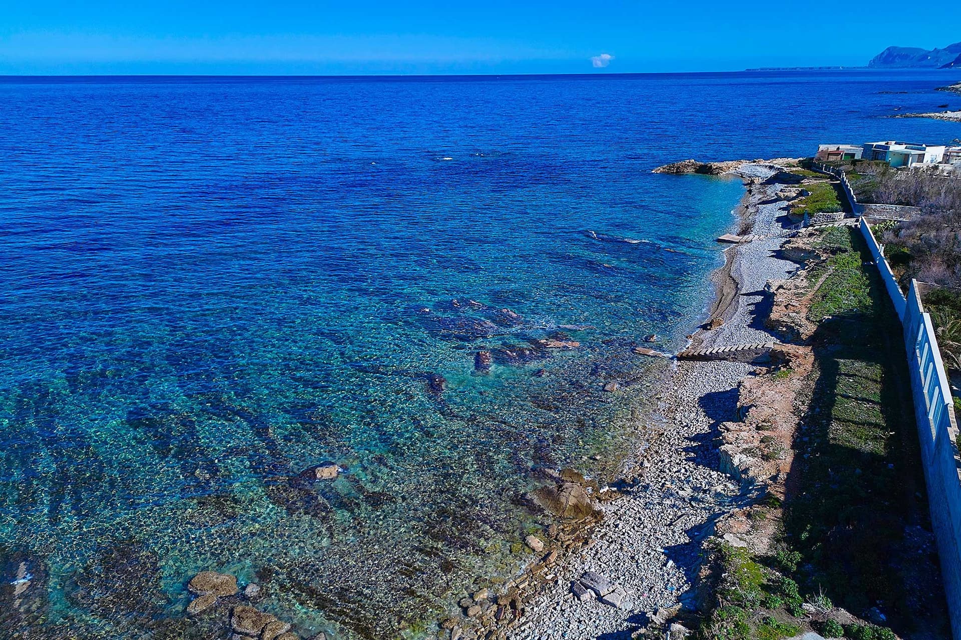 Aerial view of a rocky coastline with clear blue water, a pebble beach, and a distant mountain under a bright blue sky.