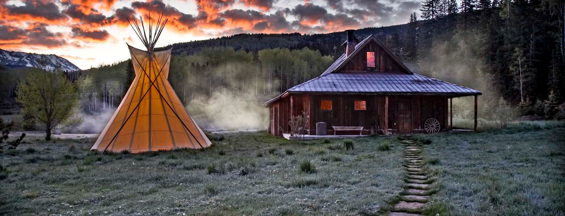 A rustic cabin and illuminated teepee at dusk, surrounded by a grassy field and trees, with a cloudy, colorful sky in the background.
