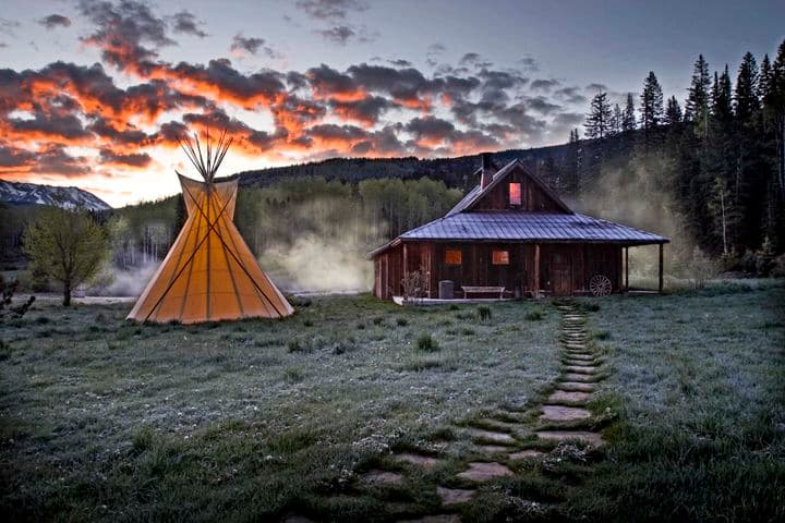 A rustic cabin and illuminated teepee at dusk, surrounded by a grassy field and trees, with a cloudy, colorful sky in the background.