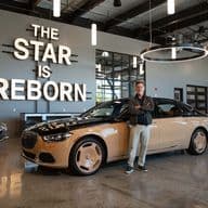 A man stands proudly in front of a two-tone Maybach sedan in a showroom. "The Star is Reborn" is lit up on the wall behind him.