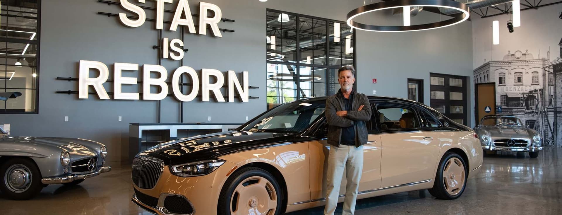 Un homme se tient fièrement devant une berline Maybach bicolore dans une salle d'exposition. "L'étoile renaît" est illuminé sur le mur derrière lui.