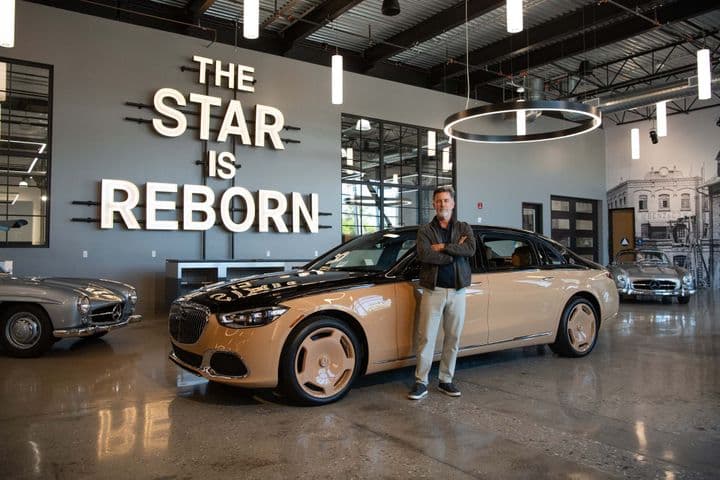 A man stands proudly in front of a two-tone Maybach sedan in a showroom. "The Star is Reborn" is lit up on the wall behind him.