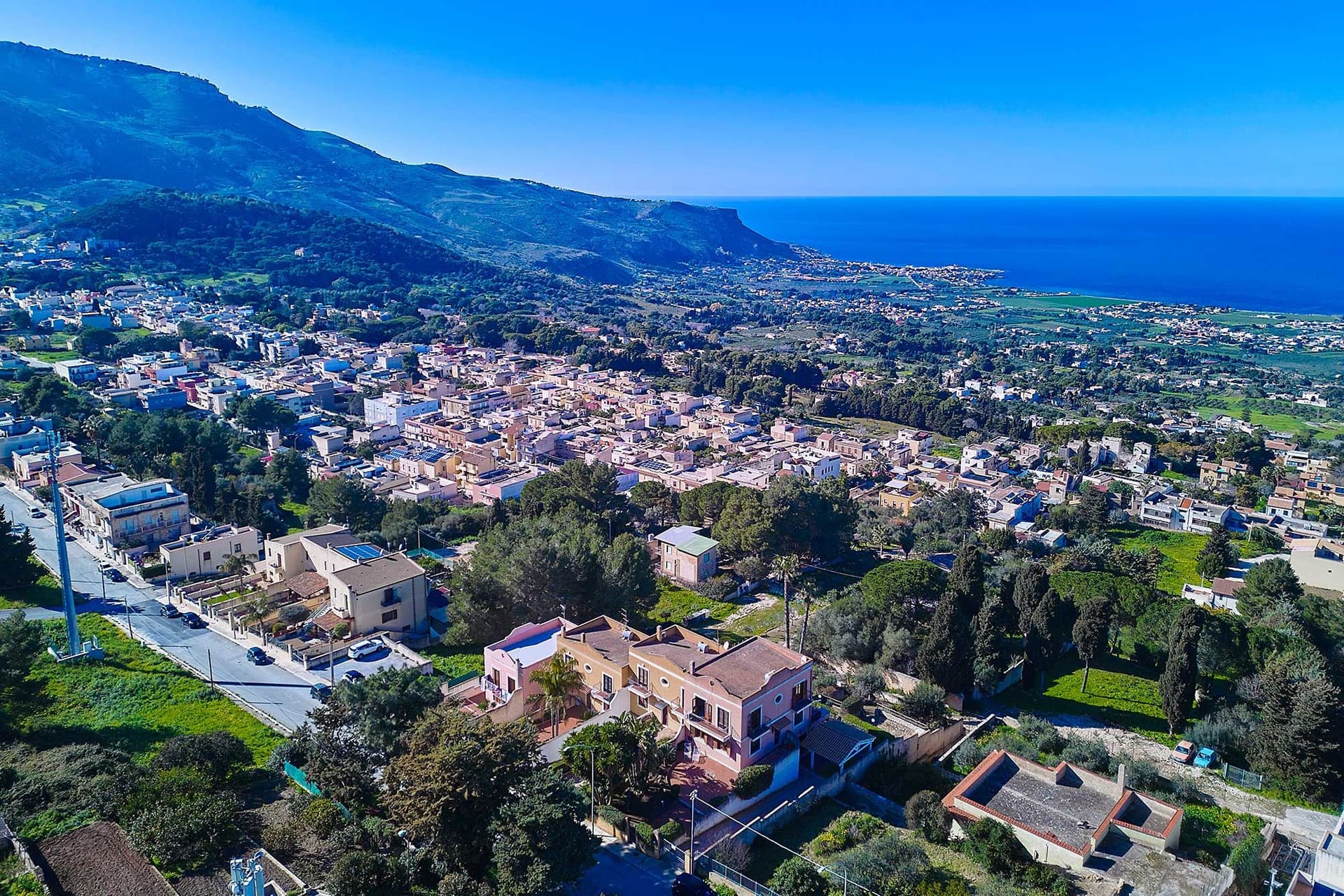 Veduta aerea di una città costiera con case a grappolo, verde lussureggiante e montagne sullo sfondo sotto un cielo azzurro e limpido.