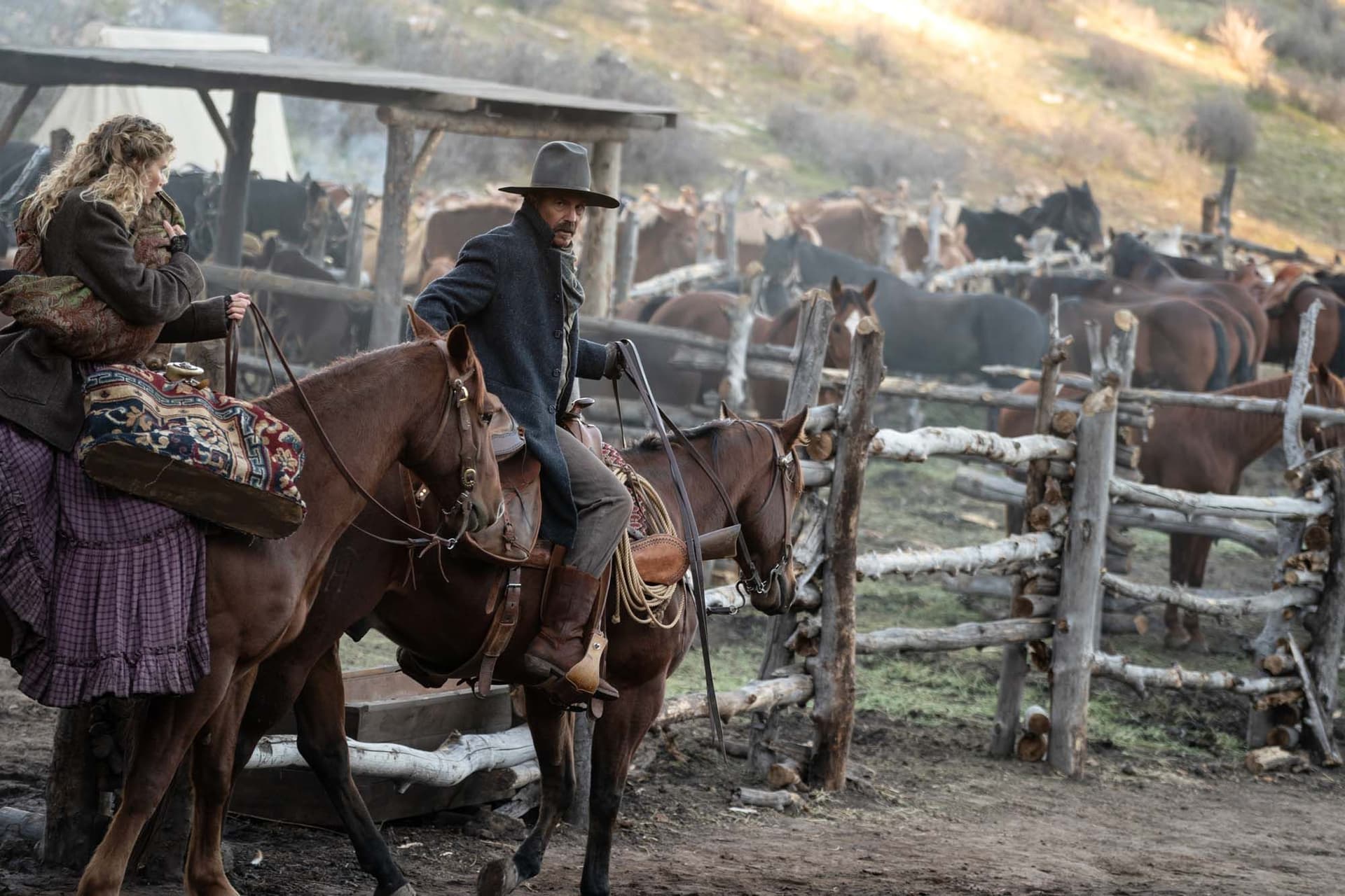 A man and woman on horseback, wearing period clothing, ride past a wooden fence with horses in the background.
