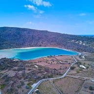 Luftaufnahme einer Küstenlandschaft mit einer kreisrunden Bucht, umgeben von Hügeln, verstreuten Häusern und kurvenreichen Straßen unter einem klaren blauen Himmel.
