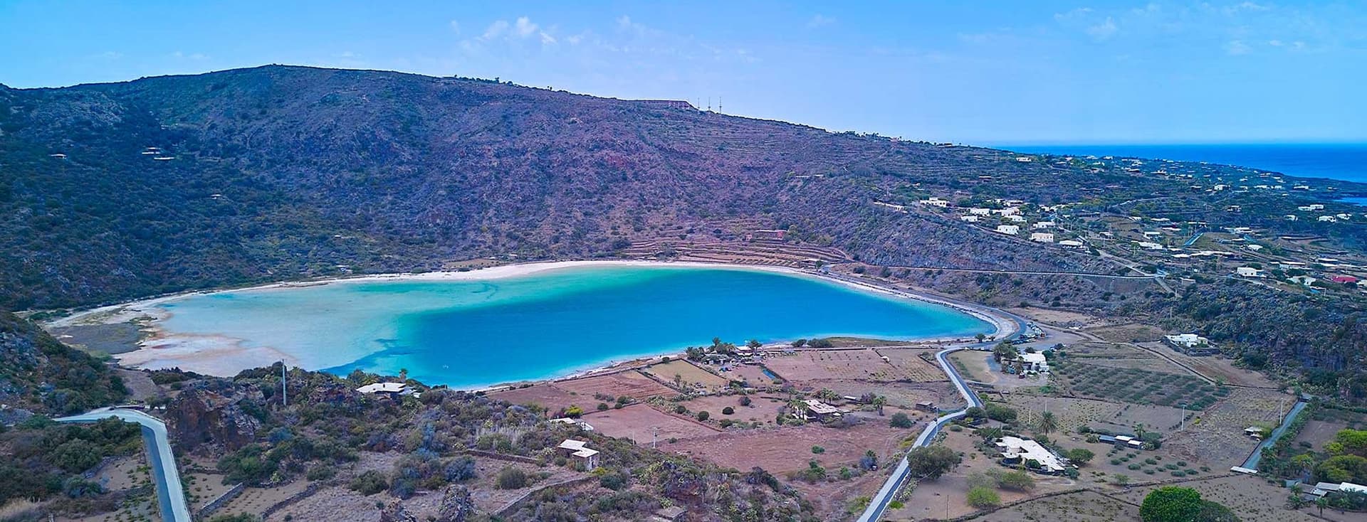 Luftaufnahme einer Küstenlandschaft mit einer kreisrunden Bucht, umgeben von Hügeln, verstreuten Häusern und kurvenreichen Straßen unter einem klaren blauen Himmel.