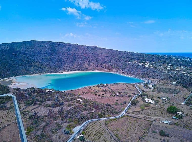 Luftaufnahme einer Küstenlandschaft mit einer kreisrunden Bucht, umgeben von Hügeln, verstreuten Häusern und kurvenreichen Straßen unter einem klaren blauen Himmel.