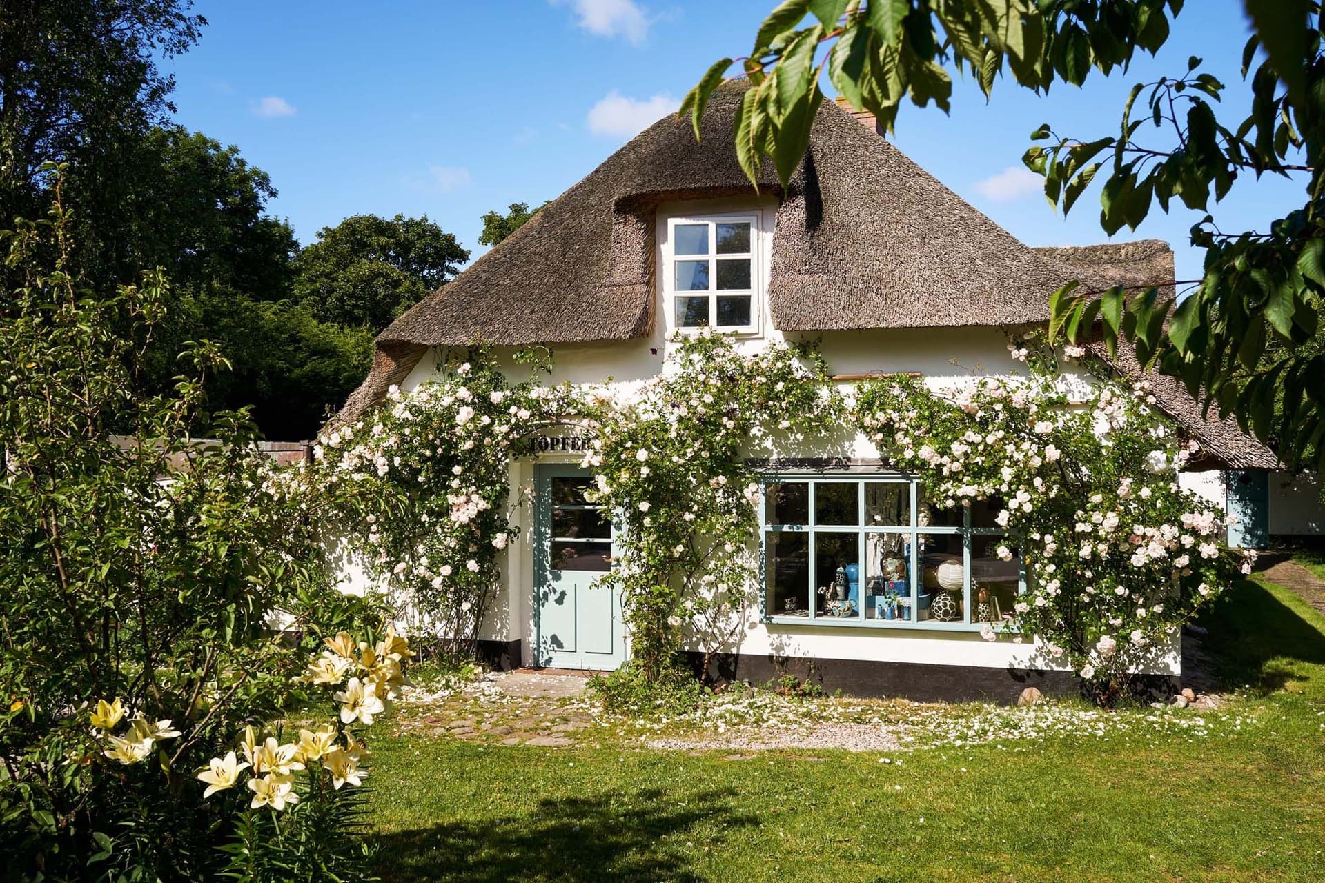 A charming cottage with a thatched roof, surrounded by lush greenery and blooming flowers under a clear blue sky.