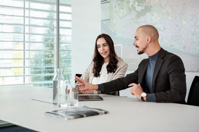 A man and woman sit at a table in a modern office, discussing while pointing at a tablet. A map is on the wall, and glasses of water are on the table.