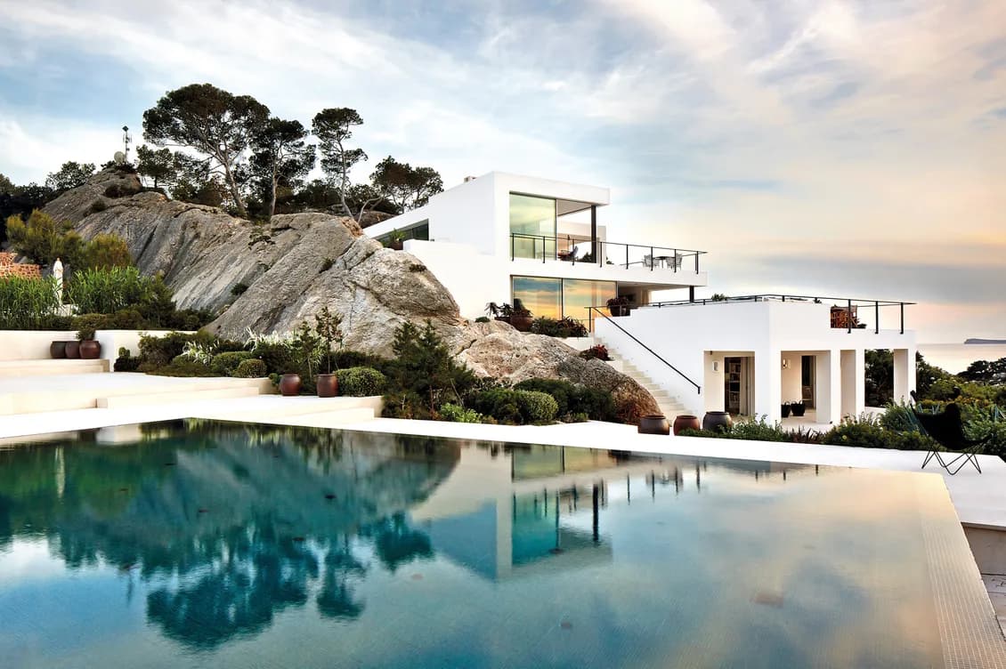 Contemporary white luxury villa built into rocky hillside with multiple levels, floor-to-ceiling windows, and an infinity pool reflecting the sky and surrounding trees. The minimalist architecture contrasts beautifully with the natural rock formations and Mediterranean vegetation, overlooking what appears to be a coastal view with soft sunset lighting.