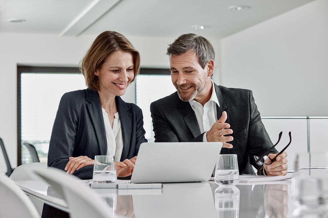 Two professionals in business attire, a woman and a man, sitting at a modern conference table reviewing content on a laptop. Both are smiling and engaged in discussion in a bright office setting with water glasses on the table.