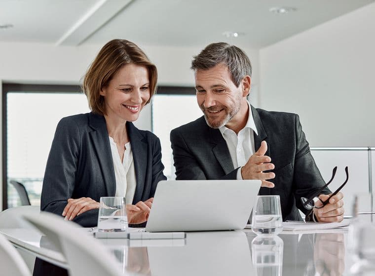 Two professionals in business attire, a woman and a man, sitting at a modern conference table reviewing content on a laptop. Both are smiling and engaged in discussion in a bright office setting with water glasses on the table.