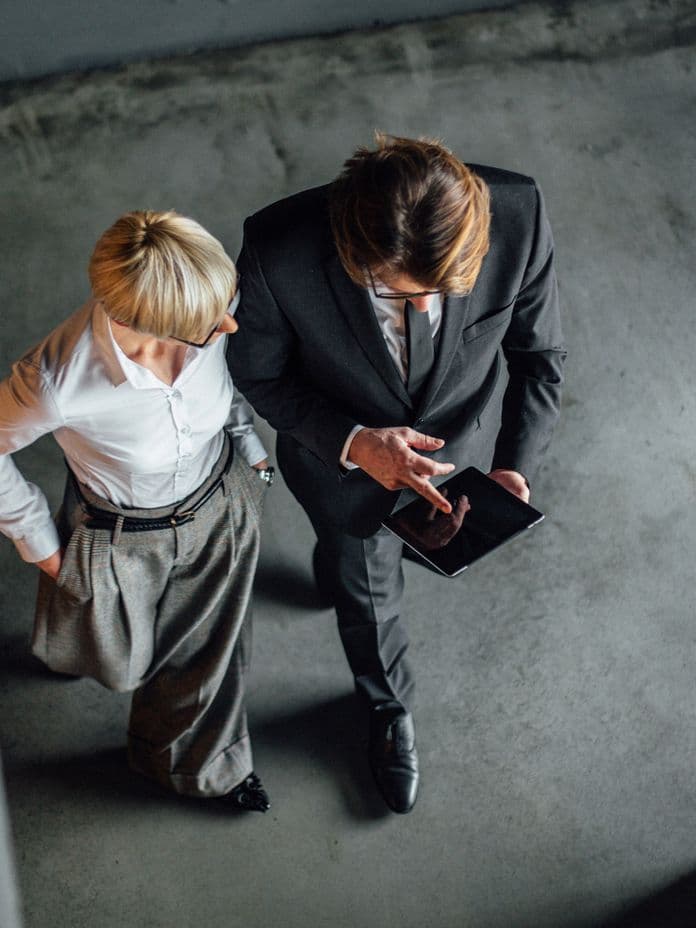 Bird's eye view of two business professionals viewed from above on a concrete floor, one wearing a black suit examining a digital device while another in a white shirt and gray pants looks on, shot in dramatic lighting with industrial style flooring.