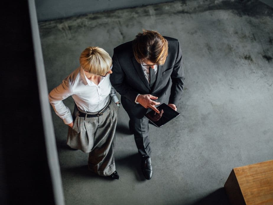 Overhead shot of two business people looking at a tablet. The man is in a suit and the woman is in a white shirt and grey pants.