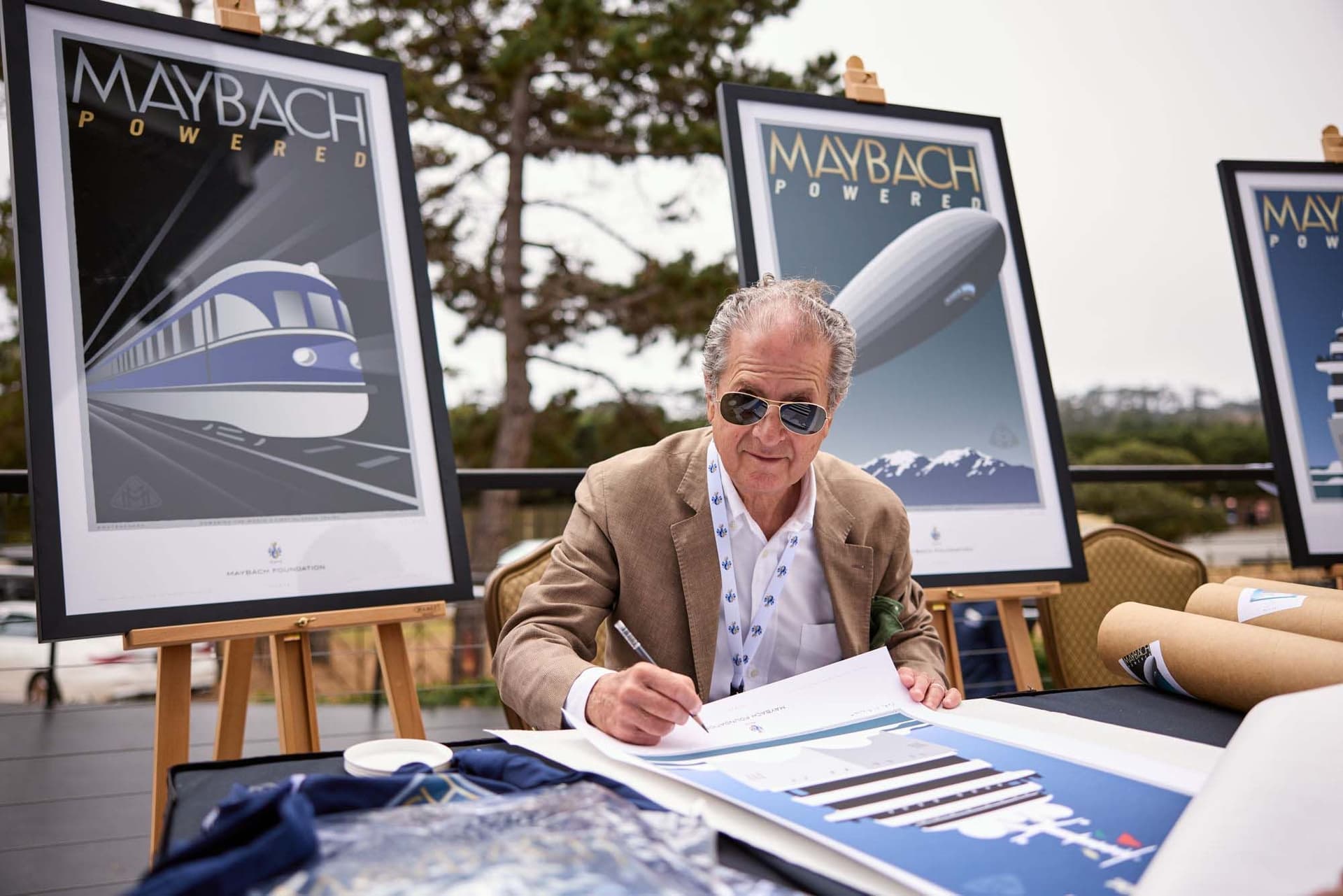 L'homme en lunettes de soleil signe une impression de la Fondation Maybach. Des affiches d'un train et d'un zeppelin sont sur des chevalets derrière lui.