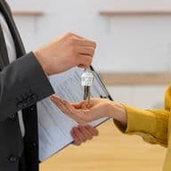 A real estate agent in a suit hands keys with a house keychain to a woman in a yellow shirt.