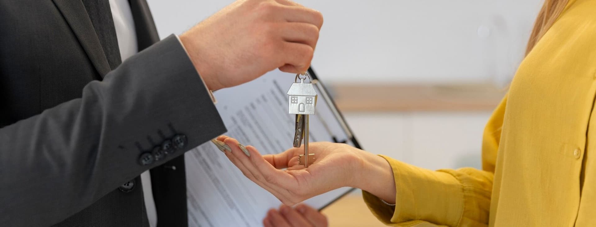 A real estate agent in a suit hands keys with a house keychain to a woman in a yellow shirt.