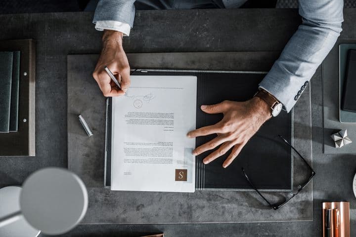 Overhead shot of a person signing a document on a gray desk. A pen, glasses, and notebooks are nearby. The person wears a gray suit.