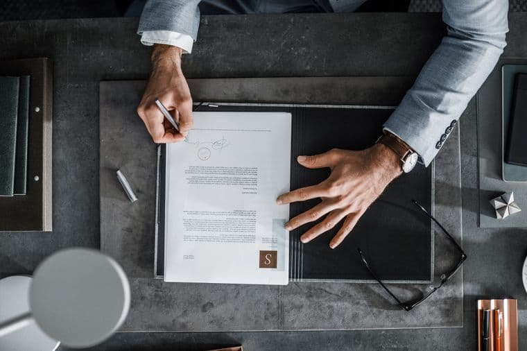 Overhead shot of a person signing a document on a gray desk. A pen, glasses, and notebooks are nearby. The person wears a gray suit.