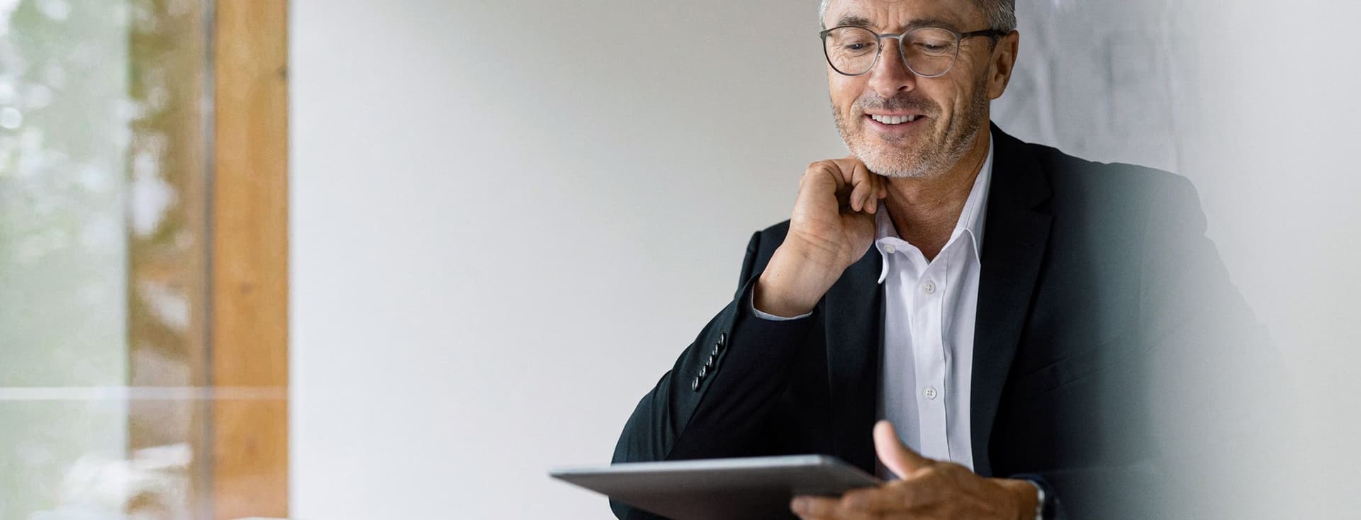 A mature businessman wearing glasses and a black suit sits at a wooden desk, smiling thoughtfully while holding a tablet. His workspace appears bright and modern with natural light coming through a window.
