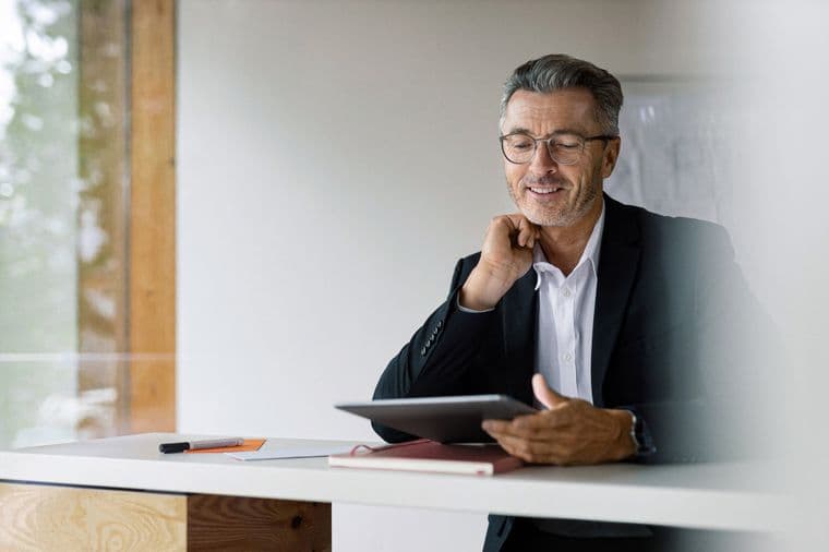 A mature businessman wearing glasses and a black suit sits at a wooden desk, smiling thoughtfully while holding a tablet. His workspace appears bright and modern with natural light coming through a window.