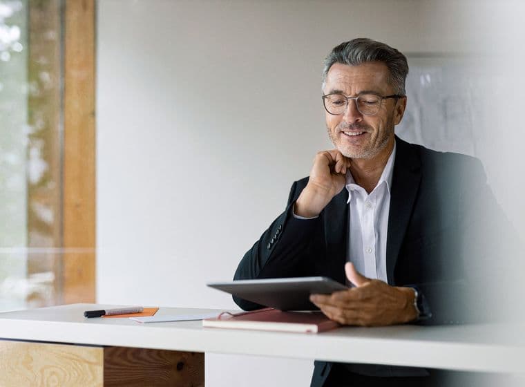 A mature businessman wearing glasses and a black suit sits at a wooden desk, smiling thoughtfully while holding a tablet. His workspace appears bright and modern with natural light coming through a window.