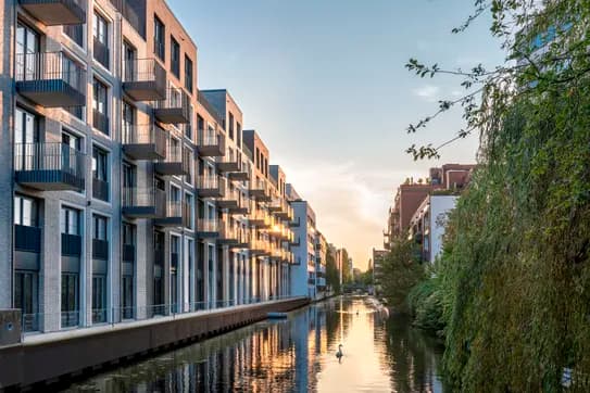 Eine Reihe moderner Backstein-Wohnhäuser mit Balkonen entlang eines Kanals; der Sonnenuntergang spiegelt sich im Wasser, während ein Schwan schwimmt, rechts sind Bäume zu sehen.