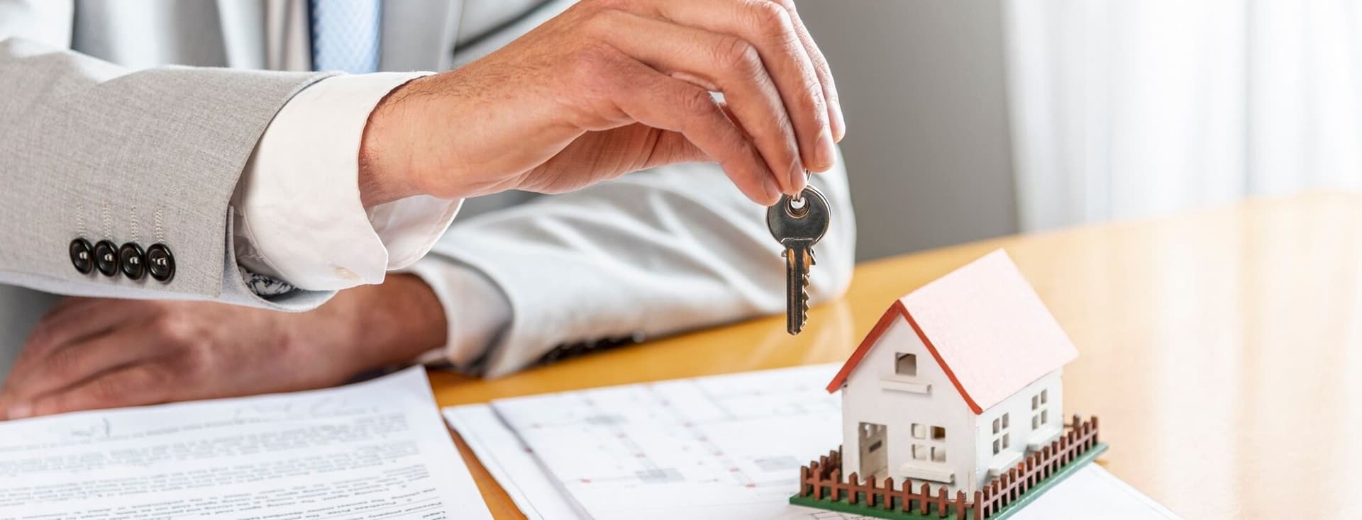 A hand in a gray suit holds a key over a miniature house on a contract.