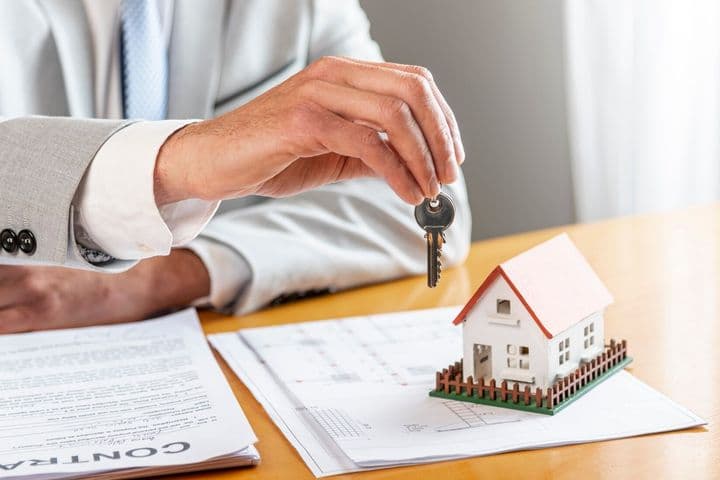 A hand in a gray suit holds a key over a miniature house on a contract.
