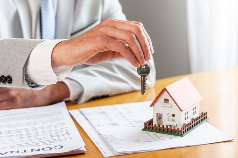 Una mano in un abito grigio tiene una chiave sopra una casa in miniatura su un contratto.