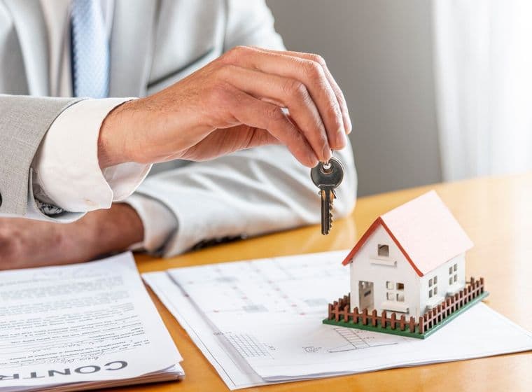A hand in a gray suit holds a key over a miniature house on a contract.