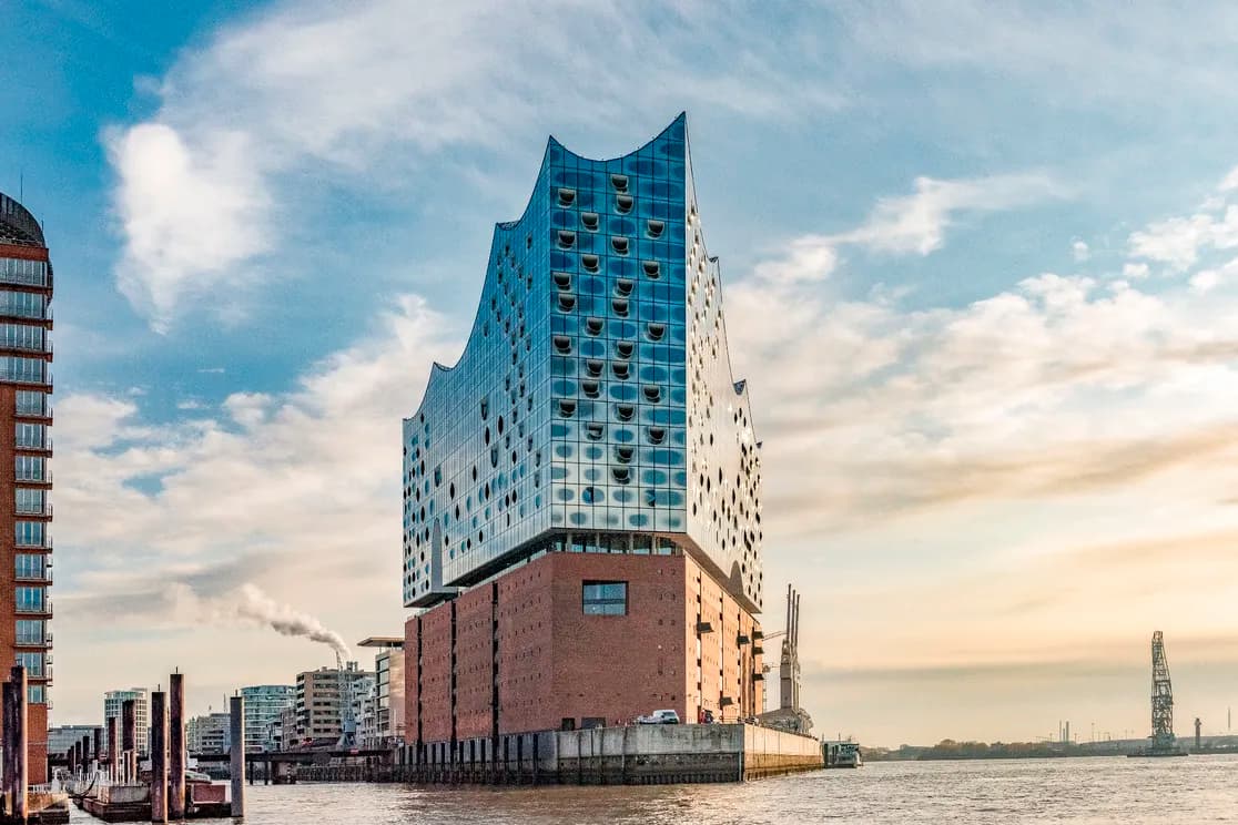 Die Elbphilharmonie in Hamburg mit einem geschwungenen Glasdach auf einem Backsteinsockel am Hafen unter blauem Himmel.