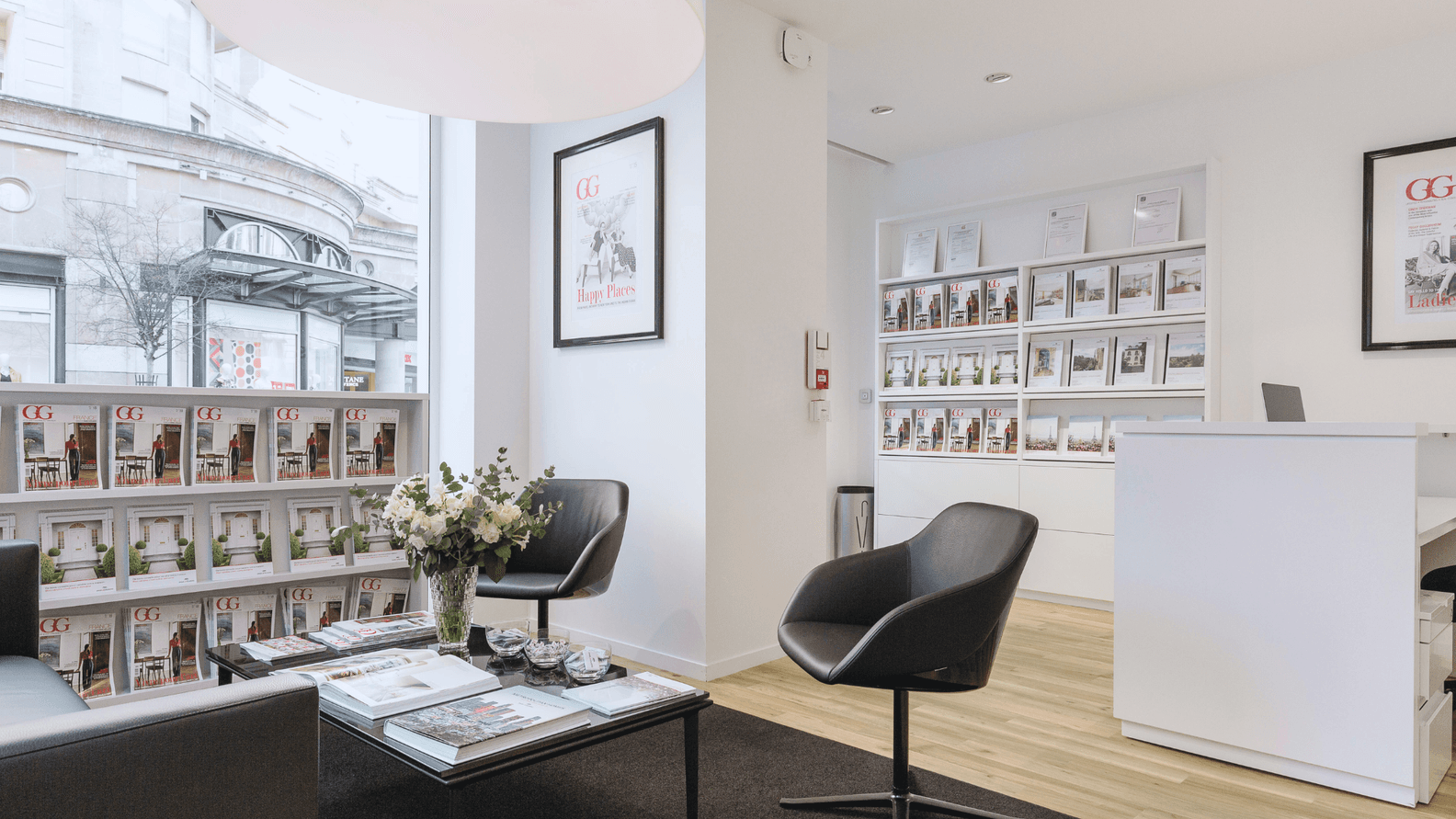 Bright real estate office with black chairs, coffee table, and magazines. A white reception desk is in the background.