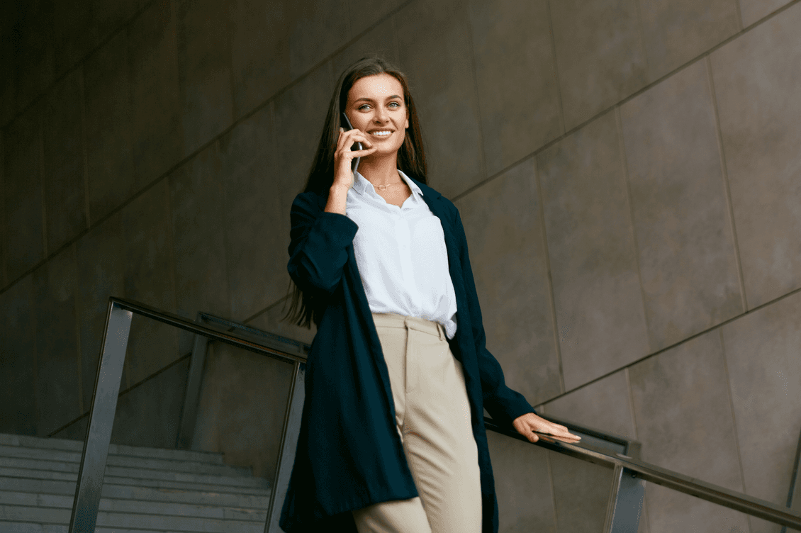 A smiling woman in a blazer and khakis walks down stairs while talking on her phone.
