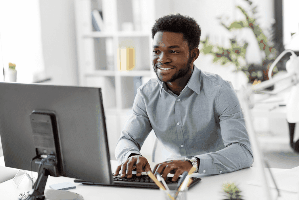 A smiling Black man in a gray shirt types on a computer in a bright, modern office.