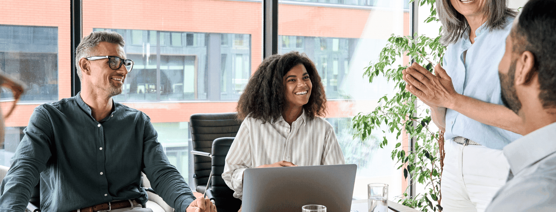 A diverse group of professionals are gathered around a conference table in a modern office, smiling and engaged in a meeting.