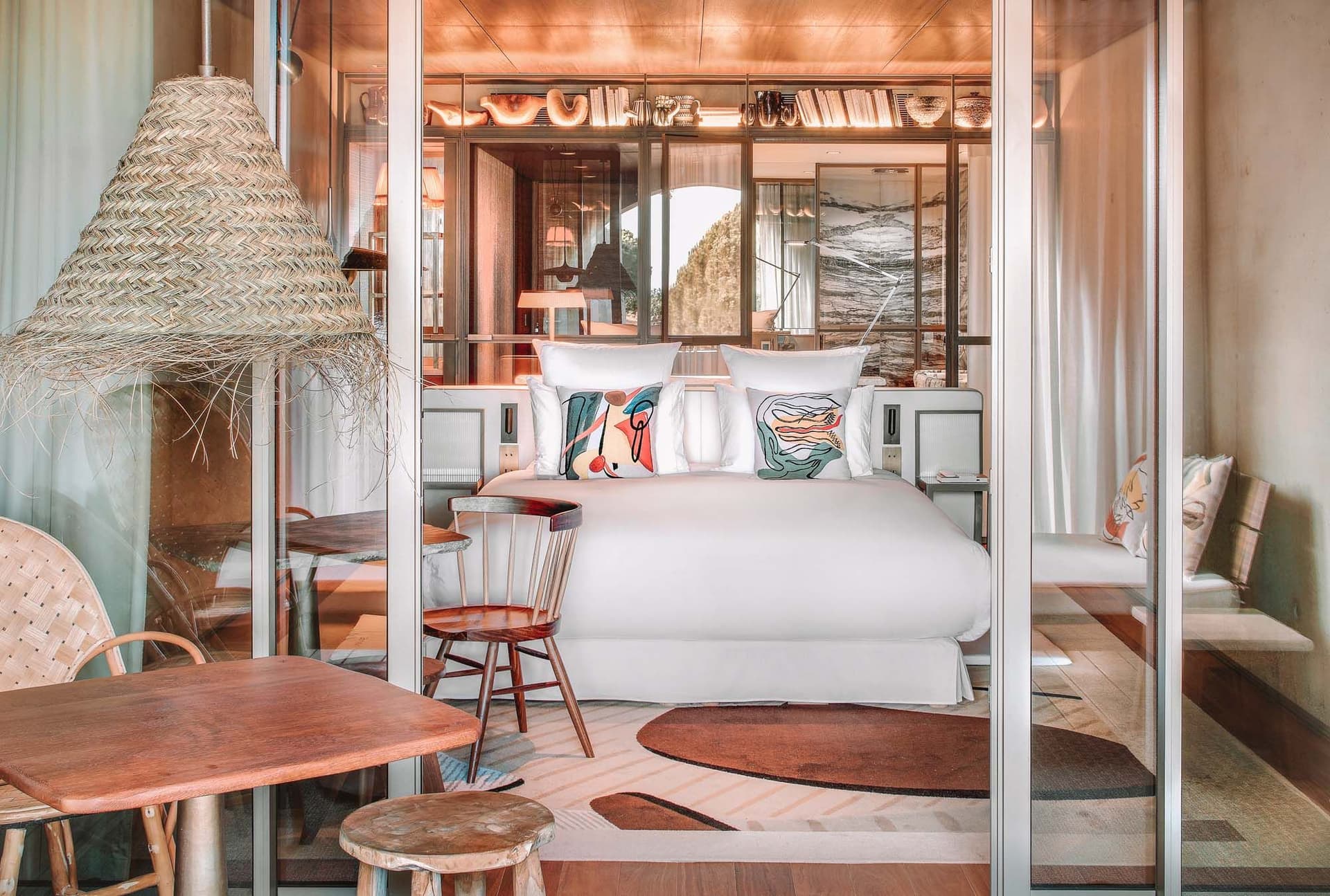 Stylish hotel room with a glass wall, cozy bed, decorative pillows, wooden furniture, and a large woven lamp, creating a warm, inviting atmosphere.