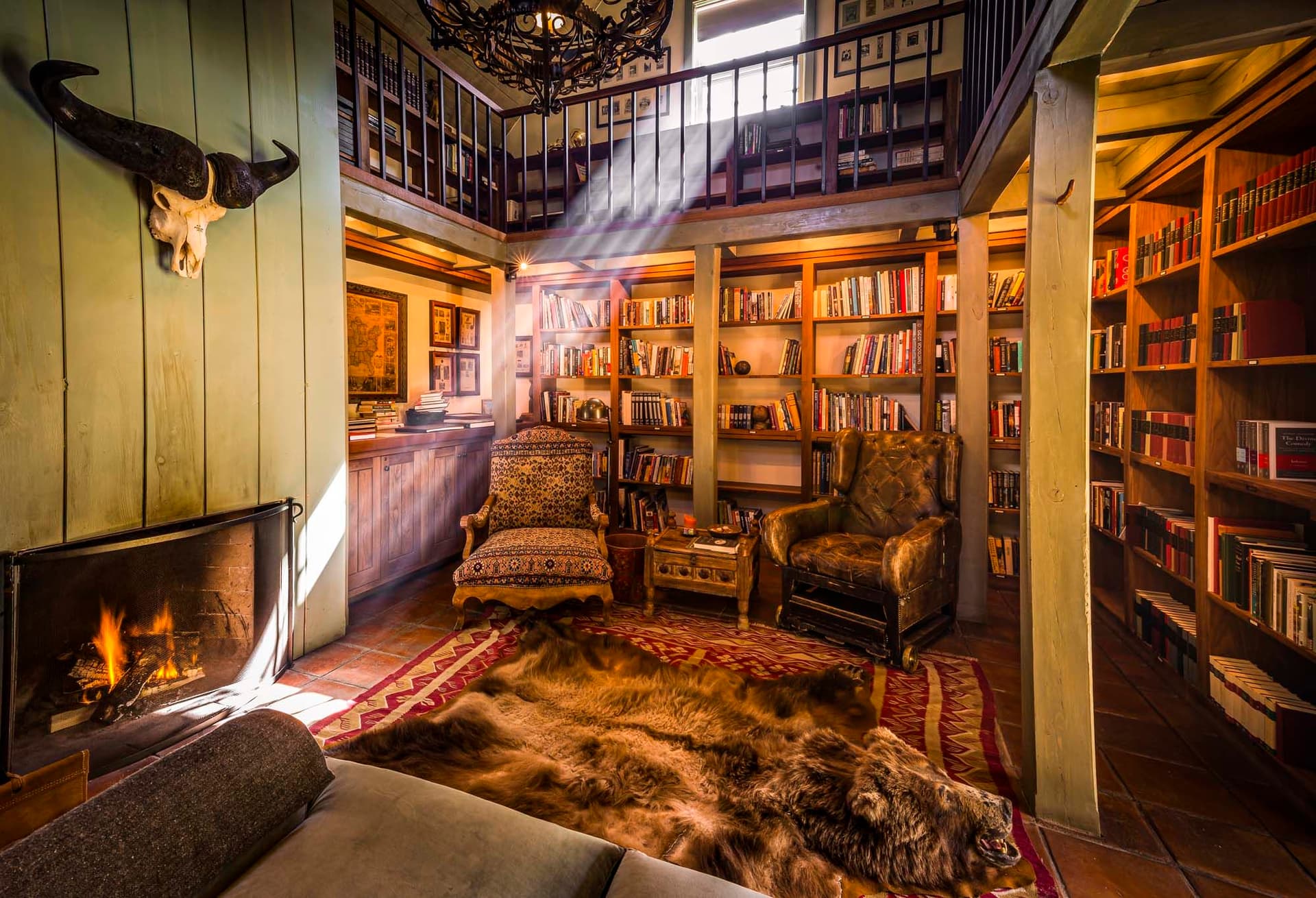 Cozy library with wooden shelves filled with books, a fireplace, animal rug, and armchairs. Sunlight streams through a window above.