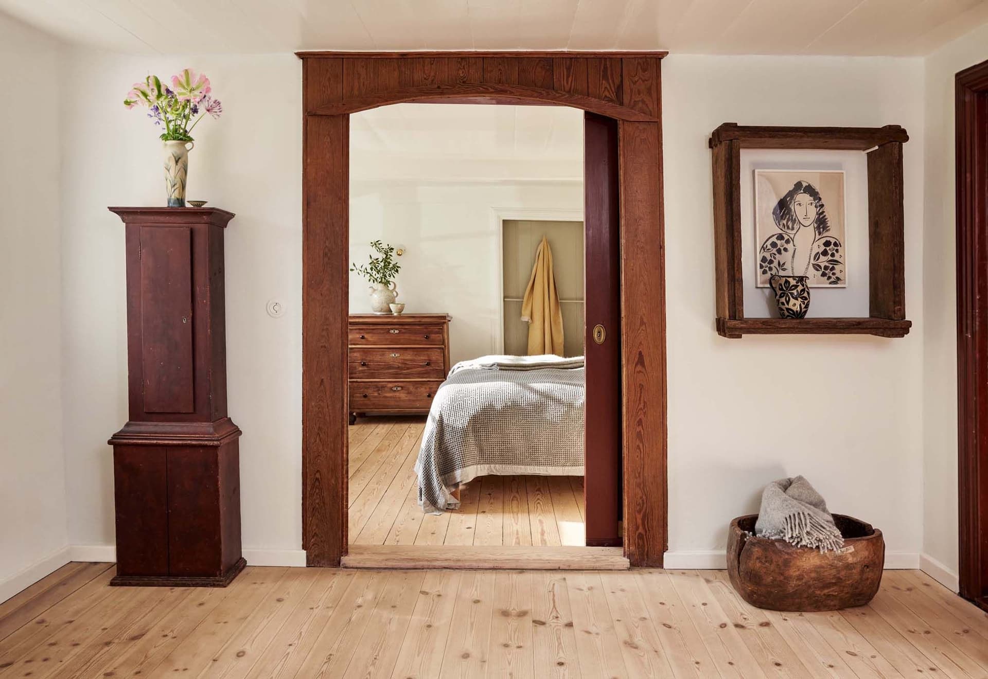 A cozy bedroom with wooden floors and furniture, featuring a bed with a gray blanket, a dresser, and a framed artwork on the wall.