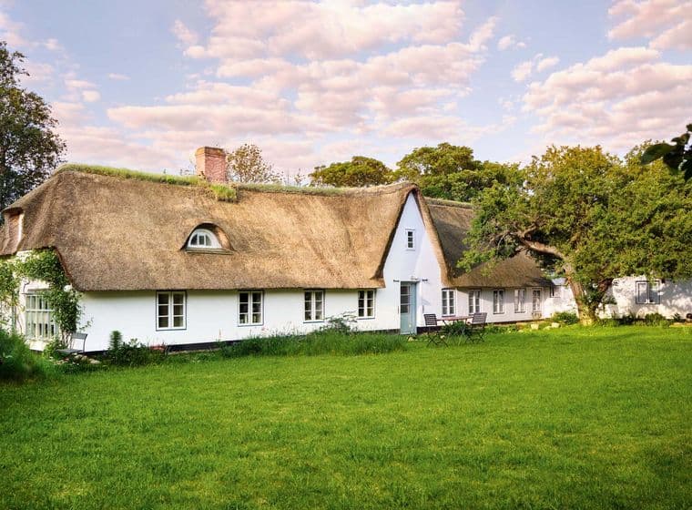 A charming white cottage with a thatched roof, surrounded by lush green grass and trees under a partly cloudy sky.