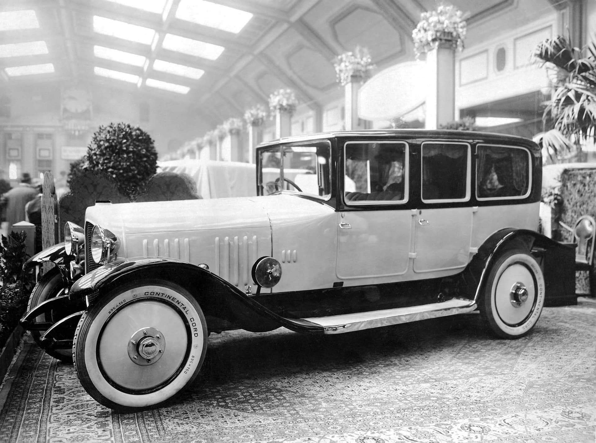 Photo en noir et blanc d'une voiture ancienne avec des pneus blancs sur un tapis orné.