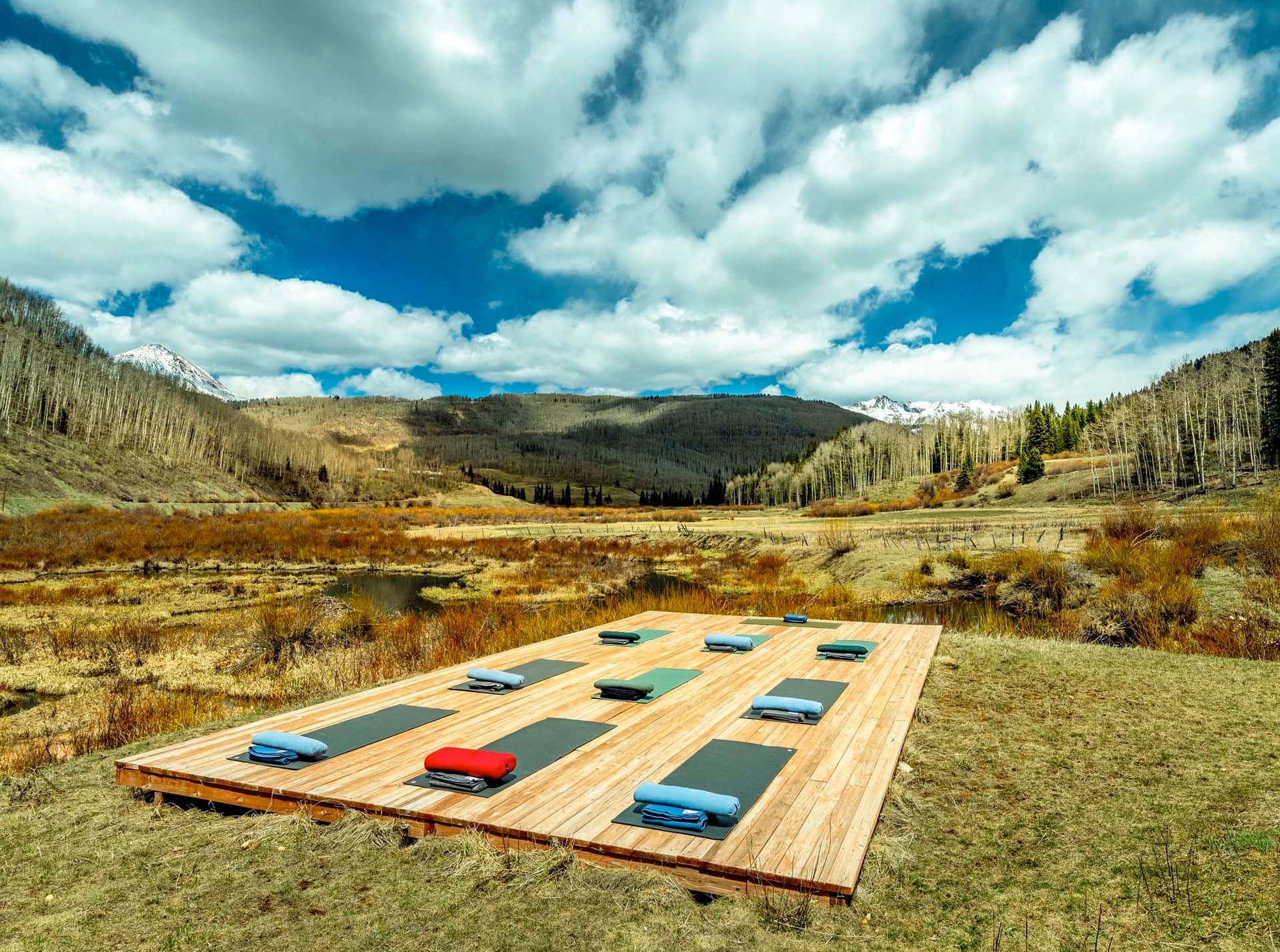 Wooden platform with yoga mats and cushions set in a scenic valley with mountains, trees, and a cloudy sky in the background.